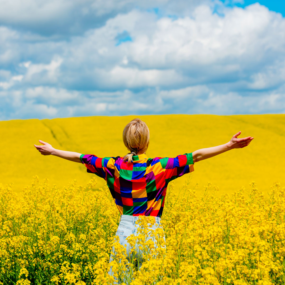 Image d'une femme dans un champs de fleurs jaune les bras ouvert. Elle symbolise l'épanouissement personnel, l'évolution et la liberté